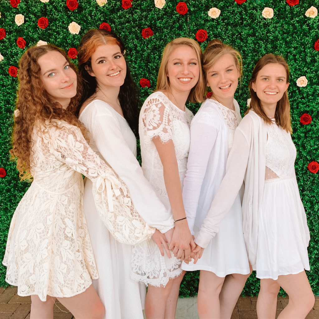 Five Alpha Gamma Delta members wearing white dresses standing in front of a flower wall. The members are holding hands and smiling.