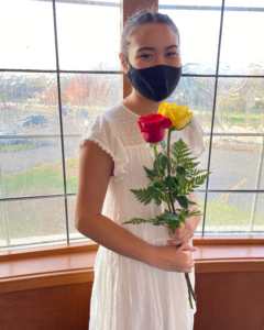 Emily Axt wearing a black mask and white dress. She is holding one red rose and one yellow rose.