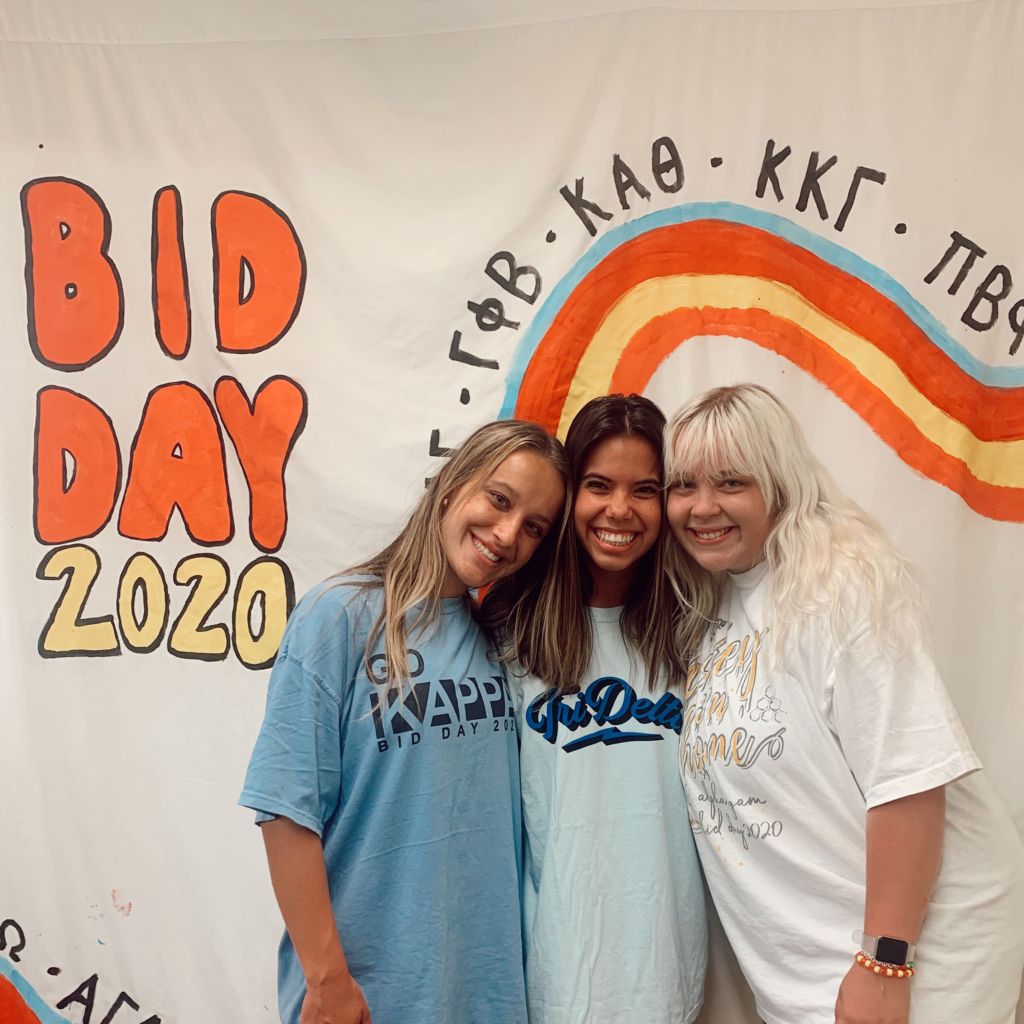 Three sorority members standing in front of a Bid Day 2020 banner.